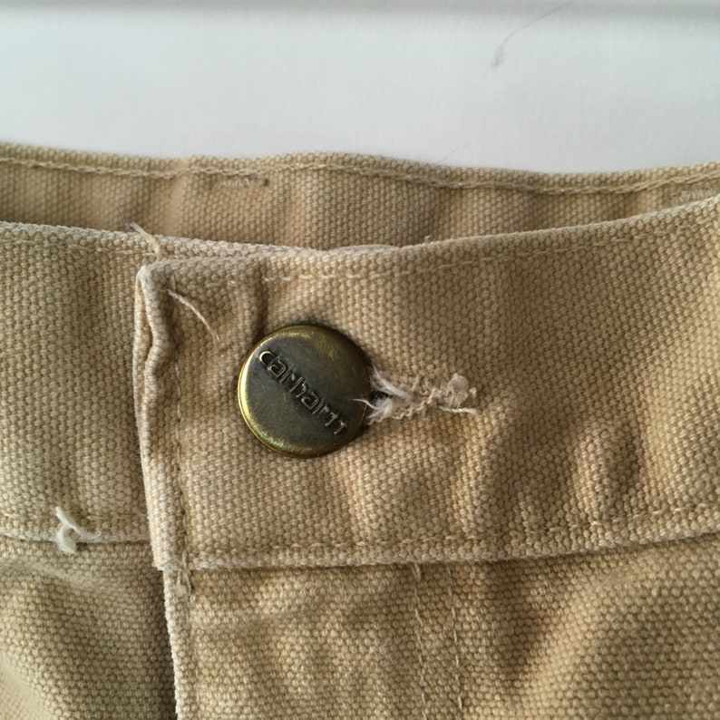 May include: Close-up of tan-colored fabric with a metal button. The button is circular with the brand name "Carhartt" embossed on it. The fabric appears to be a durable canvas material, commonly used in workwear.