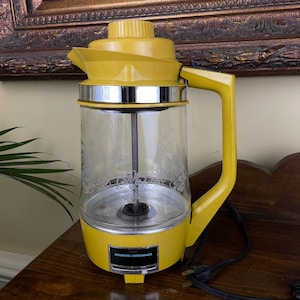 May include: A vintage yellow automatic coffee maker with a glass carafe. The carafe has a floral design. The coffee maker has a chrome band and a black power cord. The text "AUTOMATIC COFFEEMAKER" is visible on the front.