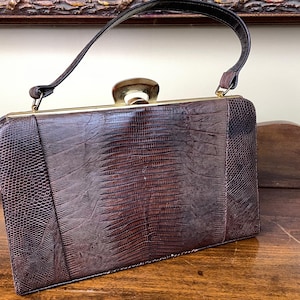 May include: A vintage brown handbag with a textured reptile-like pattern. The rectangular bag features a gold-toned clasp and trim, a black handle, and a structured design. The sides have a scaled texture, while the center has a linear pattern.