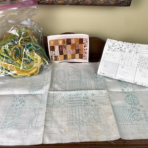 May include: A collection of embroidery supplies, including a bundle of yellow and green thread, a small framed patchwork design, and two pieces of white fabric with green stitched patterns. A pattern sheet is also included.