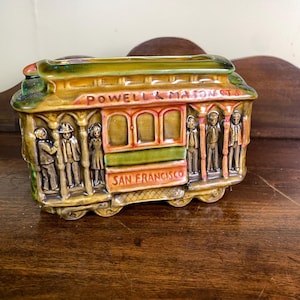 Peut inclure: Figurine de tramway de San Francisco en céramique, dans les tons marron, vert et orange. Les mots "Powell & Mason Sts" et "San Francisco" sont visibles. Les côtés présentent des personnages détaillés.