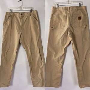 May include: Tan work pants with multiple pockets, including a hammer loop. The pants are made of a durable fabric and feature a button and zipper closure. The back of the pants has a brown leather patch with a logo.