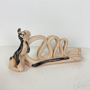 May include: A ceramic sculpture featuring a stylised dog figure with black and beige glaze. The dog is positioned next to a series of three interconnected, wavy, beige-coloured loops that extend horizontally across the base of the piece.