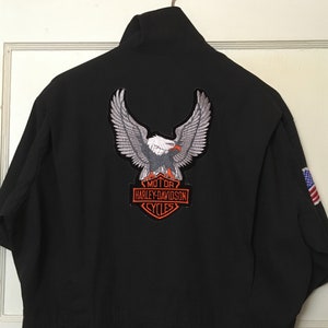 May include: Black short-sleeved jacket with a large embroidered eagle patch on the back. The patch features the Harley-Davidson logo in orange and white. An American flag patch is visible on the sleeve.
