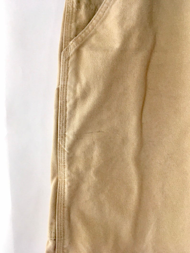 May include: Close-up of tan-colored work pants. The image shows a pocket and the seam detailing on the pants. The fabric has a textured appearance. The pants are made of a durable material.