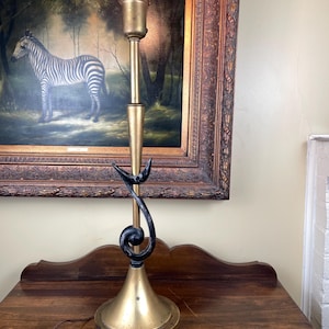 May include: A vintage brass table lamp with a black, stylized swirl design. The lamp has a conical base and a long, slender stem, topped with a lightbulb. The lamp is on a wooden surface, with a framed painting of a zebra in the background.