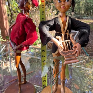 May include: Two vintage wooden dolls. One doll is dressed in a red velvet outfit with gold trim. The other doll is wearing a black outfit with gold trim and holding a harp. The dolls are approximately 16 inches tall.