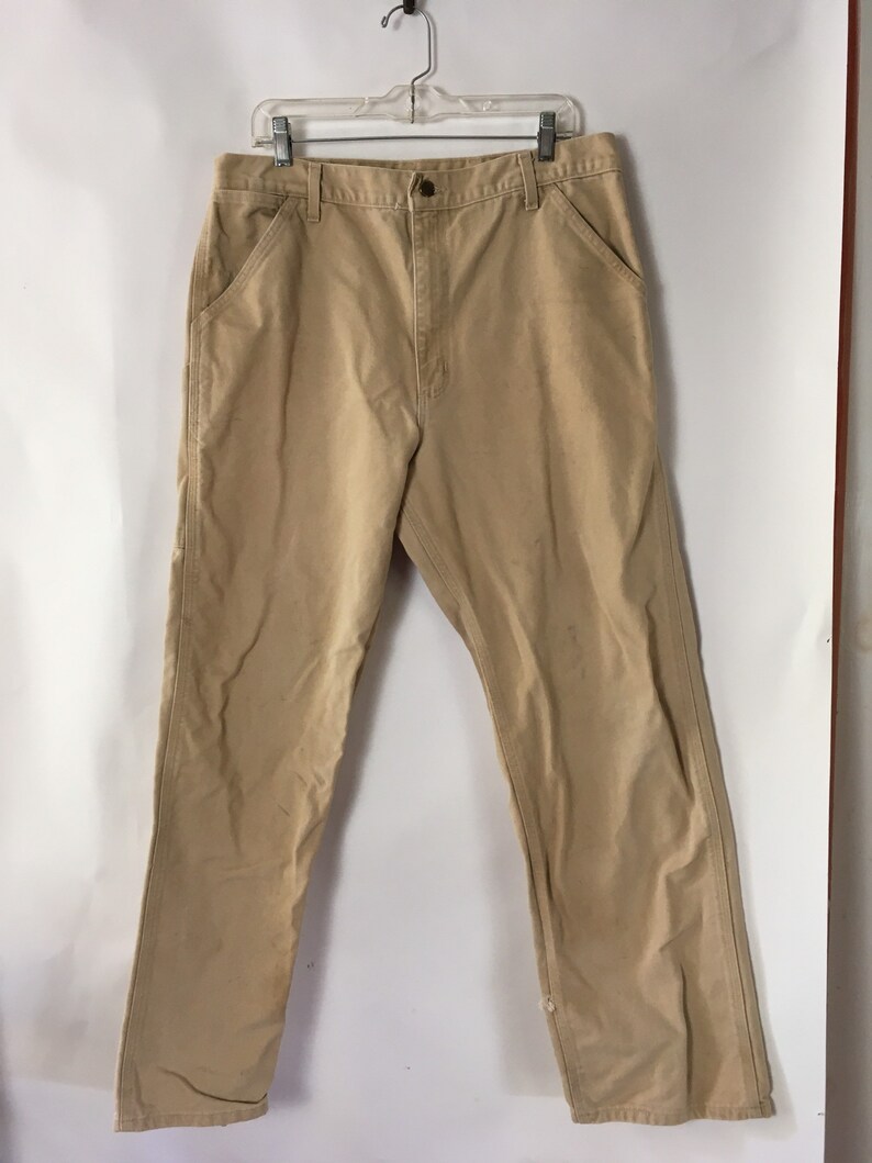 May include: Tan work pants with a button closure, front pockets, and a straight leg. The pants are hanging on a clear hanger against a white background. The pants appear to be made of a durable fabric.