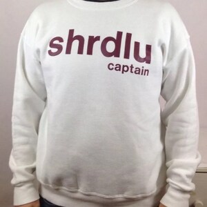 May include: White sweatshirt with the word "shrdlu" in burgundy letters above the word "captain". The sweatshirt has long sleeves and a crew neck. The person is wearing blue jeans.
