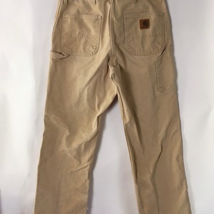 May include: Khaki-colored work pants with multiple pockets, including two back pockets and a side pocket. The pants feature a brown leather patch with a logo. The pants are hanging on a metal hanger.