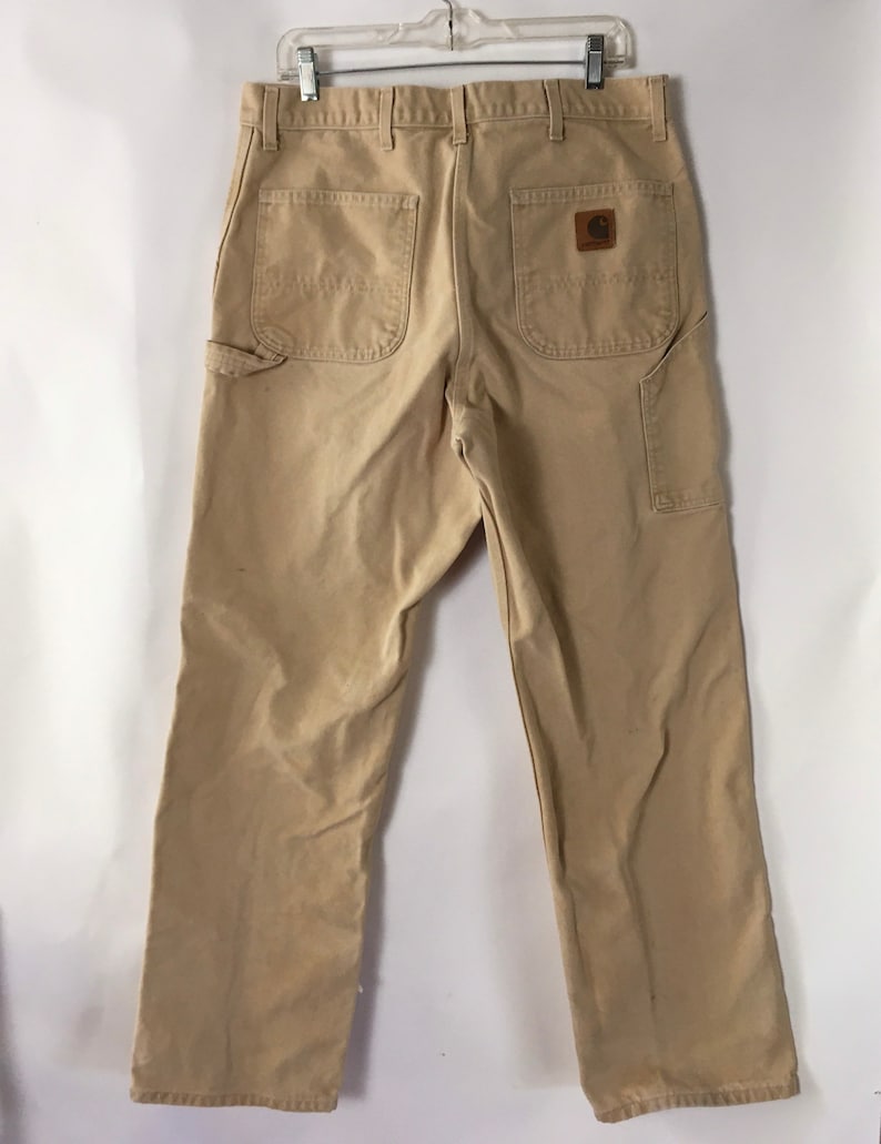 May include: Khaki-colored work pants with multiple pockets, including two back pockets and a side pocket. The pants feature a brown leather patch with a logo. The pants are hanging on a metal hanger.
