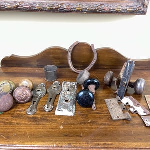 Peut inclure: Une collection de quincaillerie ancienne, comprenant des poignées de porte, des charnières et un fer à cheval, disposée sur une surface en bois. Les poignées de porte présentent des finitions en laiton, bronze et noir. La quincaillerie montre des signes d'âge et d'usure.