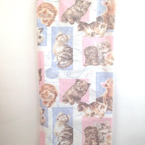 May include: A fabric panel featuring a repeating pattern of kittens in various poses. The kittens are in shades of brown, gray, and orange, set against a background of pink and blue squares. The fabric is hanging from a clear plastic hanger.