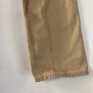 May include: Close-up of tan-colored pants leg. The fabric shows some discoloration and wear, with a visible hem at the bottom. The pants are made of a sturdy material, likely for everyday wear.