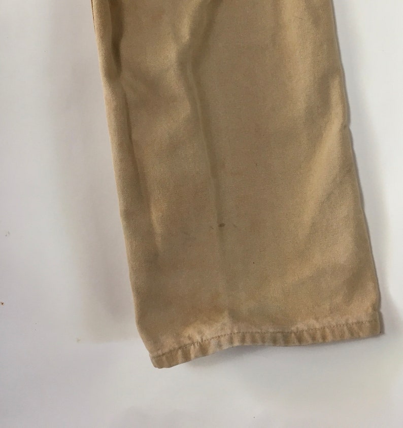 May include: Close-up of tan-colored pants leg. The fabric shows some discoloration and wear, with a visible hem at the bottom. The pants are made of a sturdy material, likely for everyday wear.