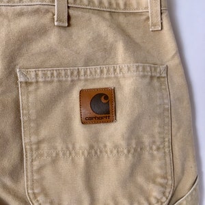 May include: Close-up of tan work pants with a back pocket and a brown leather Carhartt brand patch. The pants have belt loops and a metal clasp. The fabric appears to be a durable, heavy-duty material.
