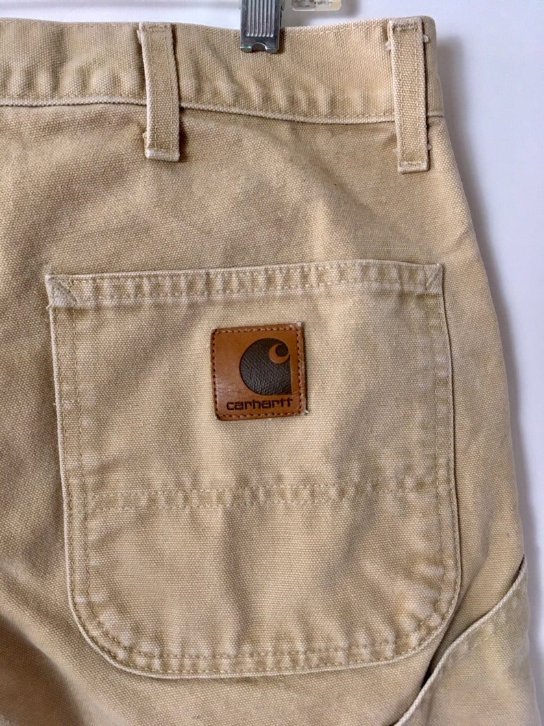 May include: Close-up of tan work pants with a back pocket and a brown leather Carhartt brand patch. The pants have belt loops and a metal clasp. The fabric appears to be a durable, heavy-duty material.