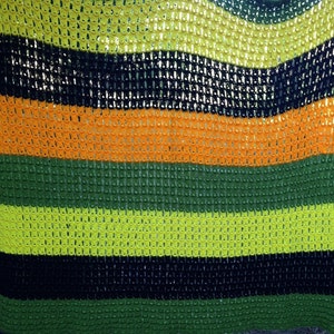 May include: A handmade, open-weave blanket with horizontal stripes in various colors. The stripes include black, orange, green, and yellow. The blanket appears to be crocheted, with a textured, grid-like pattern. The blanket is hanging outdoors.