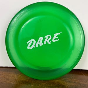 May include: A vibrant green plastic flying disc featuring the white word "DARE". The disc has a ridged outer edge and a smooth central area. This recreational toy is designed for outdoor play.