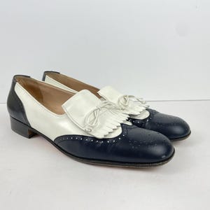 May include: A pair of two-tone loafers featuring a dark navy blue leather toe and heel counter with decorative brogue perforations. The vamp is white leather with a fringed kiltie overlay and a thin white leather bow tie detail on top.