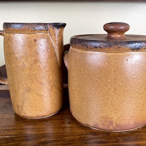 May include: A tan ceramic pitcher and a lidded jar. The pitcher has a handle and a spout. The jar has a rounded body with a lid and two small handles. Both pieces have a dark brown rim and a speckled texture.