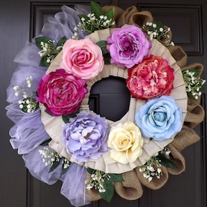 May include: A decorative wreath featuring a variety of colorful faux flowers, including pink, purple, red, and blue roses. The wreath has a burlap and pleated fabric base, with accents of white baby's breath and lavender tulle.