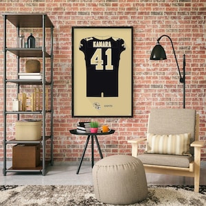 May include: A framed poster featuring a black and gold football jersey with the name "KAMARA" and the number "41". The jersey design includes the Saints logo and the word "SAINTS" at the bottom. The poster is displayed on a brick wall.