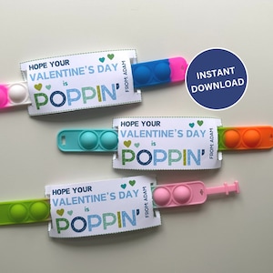 May include: A collection of colorful pop-it fidget toys with Valentine's Day cards. The cards read "Hope Your Valentine's Day is Poppin'" and include the text "From: Adam". The toys are pink, blue, green, and orange.