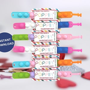 May include: A stack of Valentine's Day gifts featuring colorful pop-it toys and accompanying cards. The cards read "I HOPE YOU HAVE A POPPIN' VALENTINE'S DAY!" and "FROM MORGAN". The pop-it toys are in various colors, including pink, blue, green, and orange.