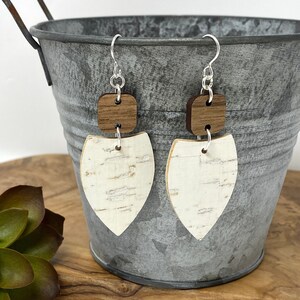 May include: A pair of handcrafted earrings featuring a square of dark wood above a white, shield-shaped piece with a textured, bark-like appearance. The earrings hang from silver-colored hooks, showcasing a natural, earthy aesthetic.