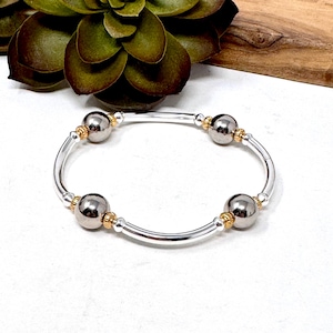 Pode incluir: Uma pulseira de prata com quatro contas cinza escuras e detalhes dourados. A pulseira é aberta e tem um design simples.