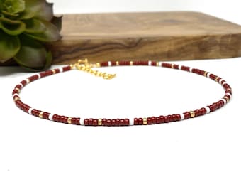 May include: A delicate choker necklace made with small red, white, and gold beads. The necklace is a simple and elegant design.