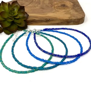 May include: Five delicate beaded necklaces in shades of blue and green. The necklaces are made of small, round beads and are strung on a silver chain. They are arranged in a graduated pattern, with the darkest blue necklace at the top and the lightest green necklace at the bottom.