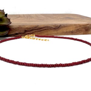 May include: A delicate, burgundy beaded necklace with a gold-colored clasp. The necklace is displayed on a white surface with a wooden board in the background. A green succulent plant is visible on the left side of the image.