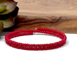 May include: A vibrant red beaded bracelet. The bracelet is made of small, round beads strung together in a spiral design. The bracelet is shown on a white surface with a wooden background and a green plant.