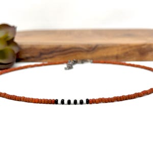 May include: A terracotta-colored beaded necklace with a silver clasp. The necklace features a row of three black beads. The necklace is displayed on a white surface with a wooden board and a succulent plant in the background.