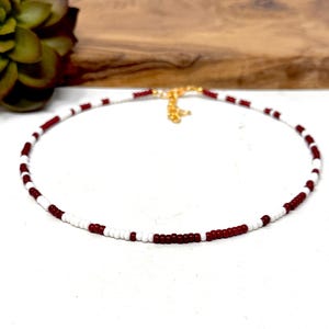 May include: A beaded choker necklace with alternating white and burgundy beads. The necklace has a gold-colored clasp and is displayed on a white surface. A succulent plant and wooden surface are in the background.