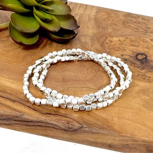 Puede incluir: Tres pulseras plateadas hechas de pequeñas cuentas cuadradas. Las pulseras están apiladas una encima de la otra sobre una superficie de madera.