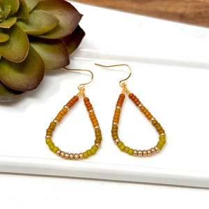May include: A pair of teardrop-shaped earrings made with small, graduated beads in shades of orange, yellow, and green. The earrings are made with gold-toned metal.