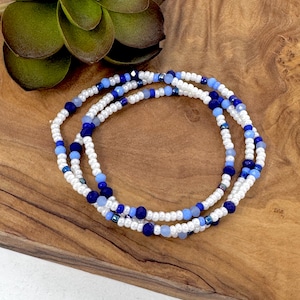 May include: Three beaded bracelets in white, light blue, and navy blue. The bracelets are made of small beads and are arranged in a circular pattern. The bracelets are on a wooden surface.