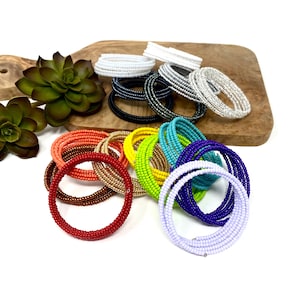 May include: A collection of colorful beaded bracelets in various shades of red, orange, yellow, green, blue, purple, and white. The bracelets are coiled and arranged on a wooden surface.