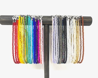 May include: A display of colorful beaded necklaces, each with a silver clasp. The necklaces are arranged in a rainbow pattern, with red, orange, yellow, green, blue, indigo, and violet beads.