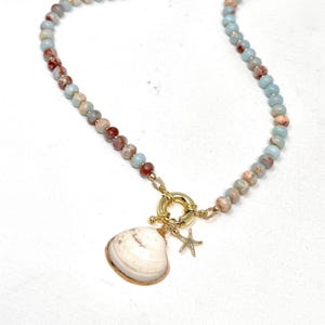 May include: A beaded necklace featuring round, multi-colored stones in shades of light blue, tan, and reddish-brown. The necklace includes a gold-toned circular clasp with a hanging white seashell pendant and a small gold-toned starfish charm.