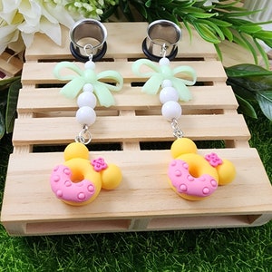 May include: A pair of silver ear gauges with a donut and bow charm. The donut charm is yellow with pink frosting and sprinkles. The bow charm is green with white beads.