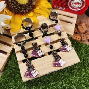 May include: A set of four silver-toned ear gauges with black bead chains and dangling charms. The charms include black cat silhouettes sitting on purple and white books, and light purple heart-shaped beads. The gauges are displayed on a wooden pallet.