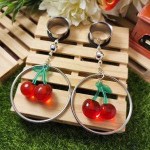 May include: A pair of silver-toned hoop earrings with dangling red cherry charms. The cherries have green stems and leaves. The earrings are displayed on a wooden surface.