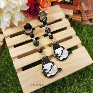 May include: A pair of black and white cat-shaped dangle earrings. The earrings feature a black cat design with a white cat inside, accented with gold trim. The earrings are connected by a chain of black beads and clear beads.
