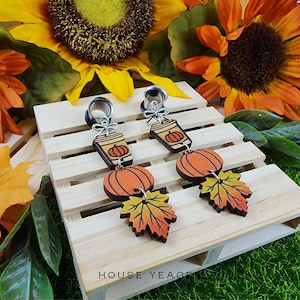 May include: A pair of silver-toned ear gauges with dangling autumn-themed charms. Each charm features a coffee cup with a pumpkin, an orange pumpkin, and a fall leaf. The earrings are set against a wooden pallet with sunflowers in the background.