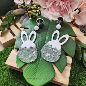 May include: A pair of white bunny-shaped earrings with clear glitter resin. The earrings are attached to silver-toned ear gauges.