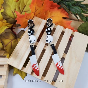 May include: A pair of black and white skull dangle earrings with a bloody knife charm. The earrings are on a wooden surface with fall leaves in the background.